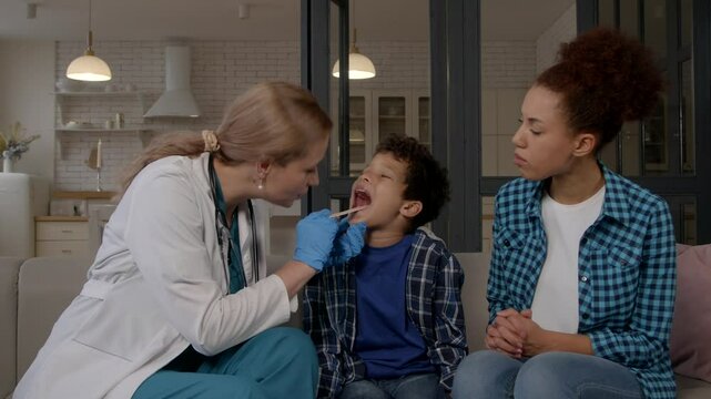 Portrait of Friendly woman ENT doctor in uniform visiting worried African American mother and sick preadolescent black son at home, doing medical checkup of child sore throat using tongue depressor.