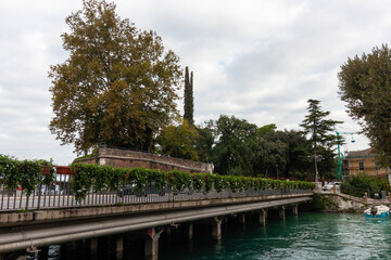 Fototapeta premium A scenic view of a bridge or embankment in Peschiera del Garda, Italy, with calm water, trees with autumn foliage, and buildings in the background