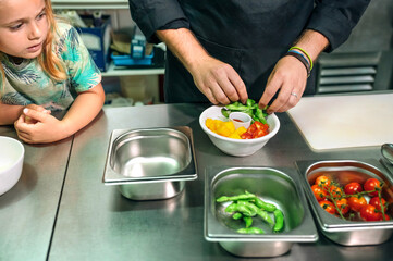 Close up of unrecognizable chef carefully preparing a vegan poke bowl while little girl watches him attentively in professional kitchen. Healthy eating and cooking education concept.