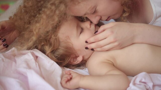 Mother with baby child lying on bed at bedroom. Beautiful mummy kissing and hug sleeping baby. Mother kissing sleeping baby. Mother caring for lovely child. Kid sleep in comfortable bed.