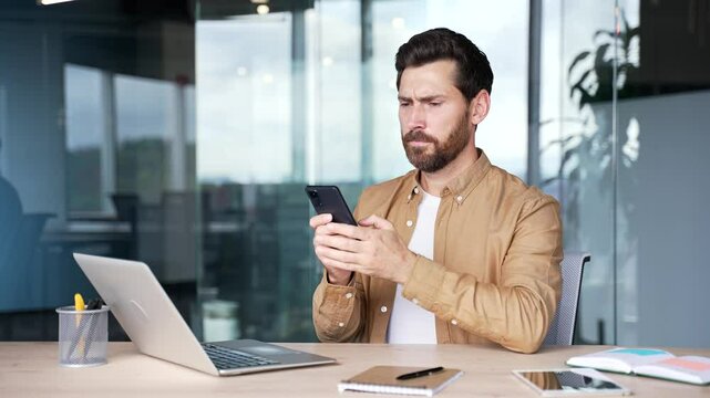 Worried businessman having problems with software or service on mobile phone sitting at workplace in modern business office. Frustrated disappointed male worker complains about bad internet connection