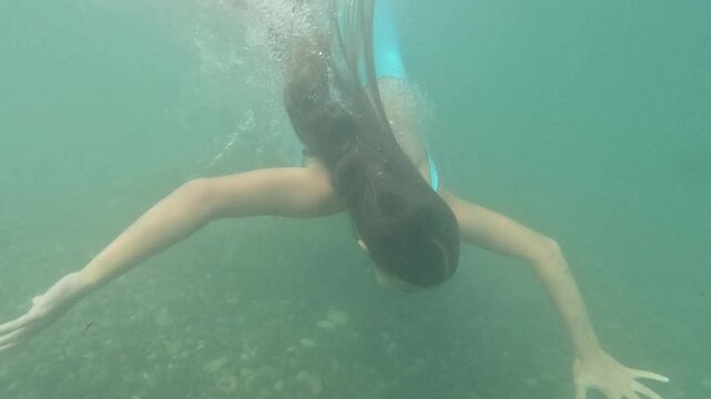 A young girl swims underwater, their body submerged with only legs and arms visible. They are swimming near the bottom, which is covered in rocks. Bubbles rise from their head as they swim. Summer