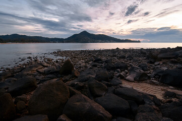 A rocky shoreline at sunset with a mountain in the background and a cloudy sky.
