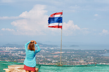 Fototapeta premium A woman in a blue jacket and red skirt takes a photo of a Thai flag on a hilltop overlooking a city and sea.