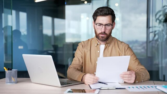 Frustrated businessman unhappy with bad financial results checking documents on laptop computer while sitting at desk in business office. Confused puzzled financier having difficulty with paper work