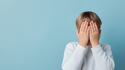 Boy with hands covering face expressing emotion in light blue background, innocence and childhood concept