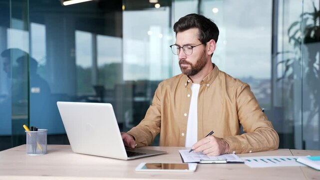 A busy confident businessman is doing paperwork using a laptop sitting in a business office. Serious worker in glasses is working performing duties, filling out documents, or making a financial report