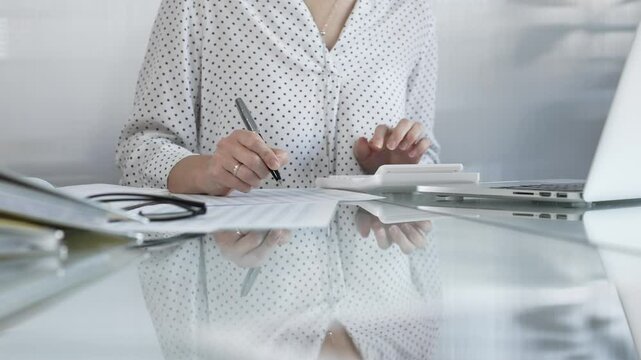 Focused businesswoman at a sleek glass desk in a contemporary office, using a calculator to crunch numbers and jotting down meticulous notes. Taxes, audit in business
