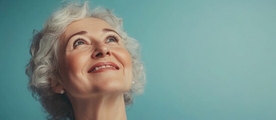 Smiling senior woman with gray curly hair gazing upwards against soft blue backdrop creating ample copyspace for text and branding concepts.
