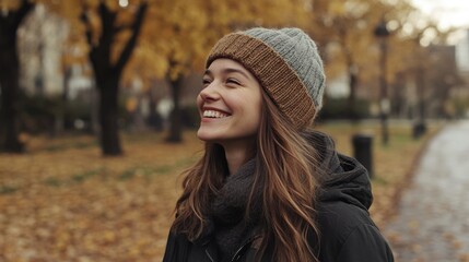 Young woman laughing in autumn park setting, wearing gray knit beanie, surrounded by fall foliage, path lined with trees, warm colors, portrait view.
