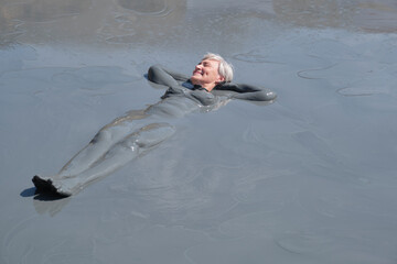 Senior woman with short gray hair relaxing in natural mud bath, fully covered in therapeutic mud