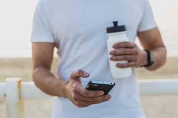 Muscular man resting on the beach promenade at sunset using his mobile phone after workout