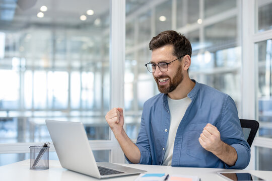 Man celebrating victory inside office at workplace, businessman working with laptop joyfully holding hands up, satisfied received excellent results achievement. - Powered by Adobe