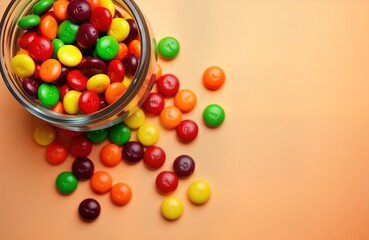 Colorful round skittles candies scattered around clear glass jar. Many-colored candies in top-down view. Background soft peach color. Delicious colorful candies make perfect treat snack for kids,