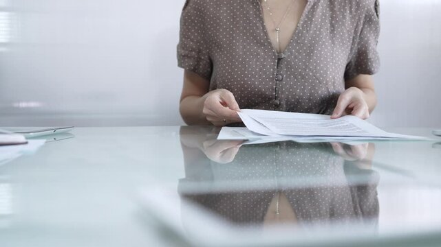 Businesswoman or accountant in brawn dotted blouse is going through paperwork while holding documents at a glass desk in a well lit, contemporary office setting. Business people concept