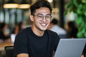 Fototapeta premium Asian young adult male working on laptop in office setting, smiling and wearing glasses Laugh at Work Week
