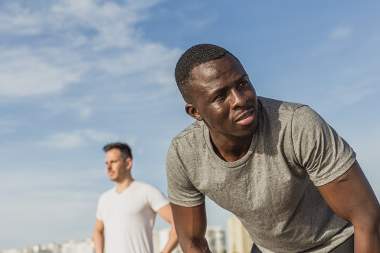 Two men, one Caucasian and one Black, stretching together before a workout on the beach at sunset