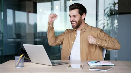 Happy excited handsome businessman received great news on laptop while sitting at workplace in business office. Smiling shocked surprised bearded entrepreneur reads a positive good message on computer