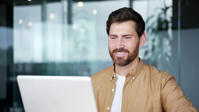 Happy handsome businessman working on a laptop computer sitting at workplace in business office. Smiling bearded male worker, banking, texting a client, chatting online. busy with a project. Close up