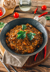 Traditional Georgian dish Ostri in a black bowl with meat pieces, vegetables, and herbs, surrounded by ingredients on rustic wooden table. Selective focus
