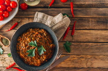 Traditional Georgian dish Ostri in a black bowl with meat pieces, vegetables, and herbs, surrounded by ingredients on rustic wooden table. Top view