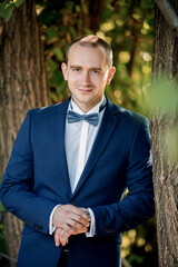 A man in a blue suit and bow tie stands next to a tree. He is smiling and looking at the camera