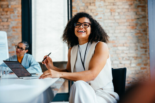 Confident young professional woman smiling in a modern office setting using laptop and pen during a business meeting