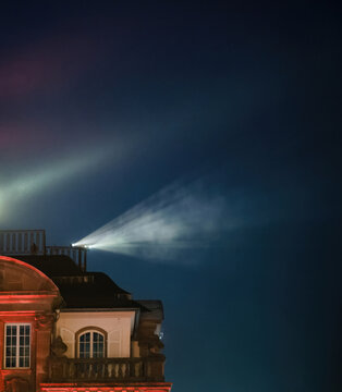 Illuminated stage lights and projected beams create an ethereal glow against a historical building, blending architectural beauty with artistic lighting effects.