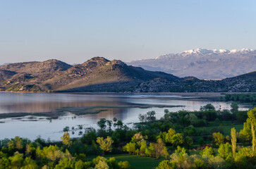 A serene landscape of Skadar Lake in Montenegro surrounded by mountains. The water is calm, reflecting the sky and the surrounding hills creating a peaceful atmosphere. 