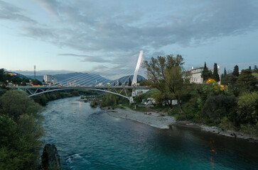 Modern Millennium Bridge in Podgorica, Montenegro, spanning the Morača River under a dramatic sky. Urban landscape with river and greenery.