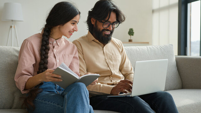 Diverse couple on couch with laptop pay bills loan payments planning household budget arabian woman wife read notes in notebook indian man husband typing data email on computer family business at home