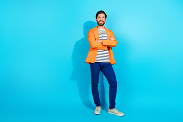 Confident man in bright orange jacket standing with crossed arms against teal background,...