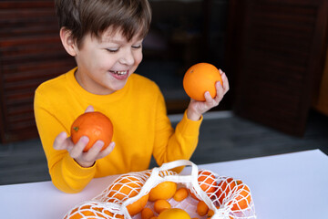 A child with a smile holds an orange and a mandarin, looking at a bag filled with citrus fruits. 
