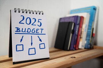 otwarty notes na półce z kartką na której zapisany jest 2025, budget oraz poniżej trzy puste pola, do których prowadzą strzałki © siwyk