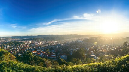 Obraz premium Golden Hour Cityscape: Panoramic view of a city bathed in the warm glow of the setting sun, nestled amidst lush green hills. A breathtaking vista of urban sprawl meeting nature's beauty. 
