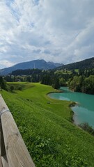 A turquoise river flows through green trees, with a small town and misty mountain peaks in the background. Pastoral Italian mountain landscape. Soraga di Fassa Dolomiti, Italy