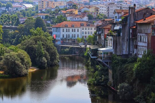 A European townscape with a river flowing through it, featuring old buildings with red rooftops and lush green trees. Amarante, Portugal.