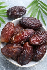 Many tasty dried dates in bowl and leaves on gray textured table, flat lay