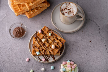 photo Delicious waffles drizzled with rich chocolate sauce, topped with colorful mini marshmallows and accompanied by a steaming cup of cappuccino on a cozy breakfast table