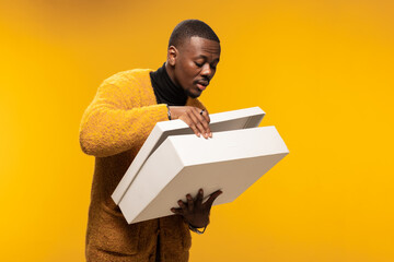Man in cozy sweater opening white gift box with curiosity in studio with golden background