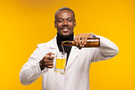Smiling man in a lab coat pours a refreshing bottle of beer into a glass mug with a cheerful expression on a yellow background