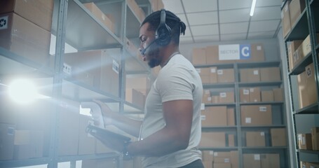 Advanced Warehouse: African American Manager Wearing Headset Scans Parcels, Checks Inventory on Tablet Computer. Worker in Full Body Powered Exoskeleton with Cardboard Box Walks in the Background.