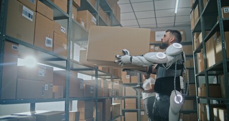 High-Tech Advanced Warehouse: Sorting Center Worker Wearing Futuristic Full Body Powered Exoskeleton Places Cardboard Parcel on Shelf. African American Manager Checks Inventory Using Tablet Computer.