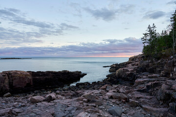 A beautiful serene evening in Acadia National Park. The blue ocean is calm, and the sky is mostly blue, with a little pink in the clouds. The rocky granite coastline in the sunset.