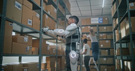 High-Tech Advanced Warehouse: Sorting Center Worker Wearing Futuristic Full Body Powered Exoskeleton Places Cardboard Parcel on Shelf. African American Manager Checks Inventory Using Tablet Computer.