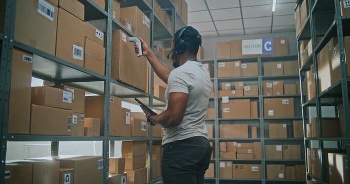 High-Tech Modern Warehouse: Worker Wearing Futuristic Full Body Robotic Exoskeleton Puts Cardboard Box on Shelf. African American Logistics Specialist Scans Parcel, Checks Inventory on Tablet Computer