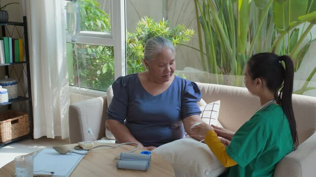 Young female medical worker putting bandage on arm of Asian senior woman while preparing her for IV drip infusion at home