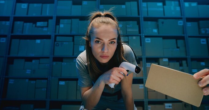 Tired Female Warehouse Employee Looking at Camera, Scanning Cardboard Boxes Using Scanner. Postal Service, Logistics Center with Shelves full of Packages in Background. Blue Neon Lighting. Portrait.