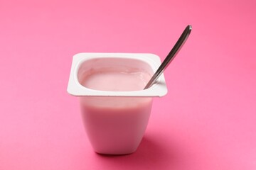Delicious yogurt in plastic cup with spoon on pink background, closeup