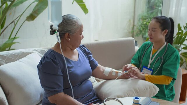 Asian young nurse putting bandage on senior patients arm while giving her intravenous vitamin course infusion during home visit